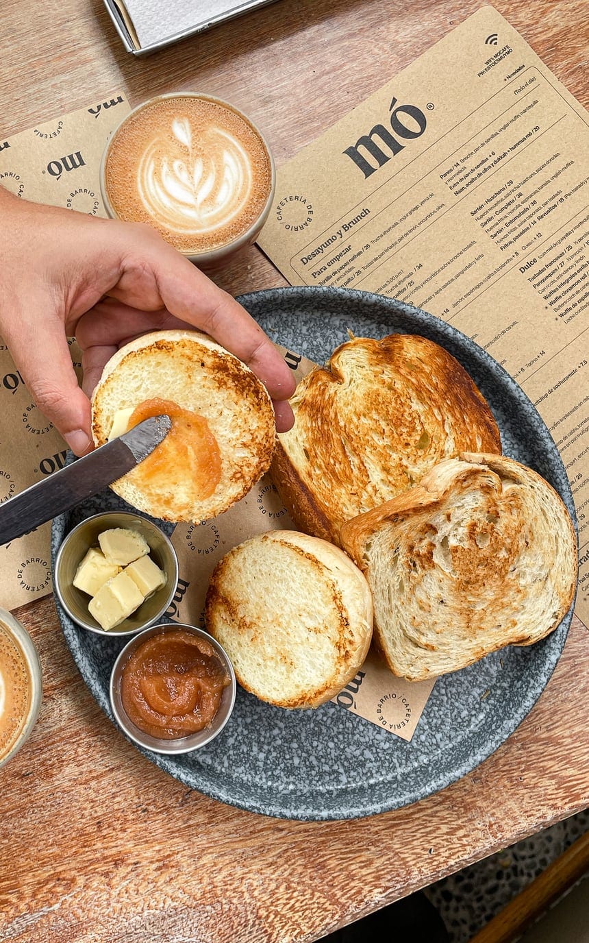 Canasta de pan con brioche, english muffin, mantequilla y mermelada sobre la carta de mó, junto a un café